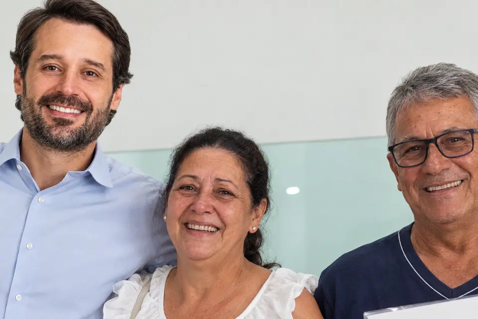 Guilherme Piai, de camisa social azul clara, sorri abraçado a uma mulher de blusa branca e a um homem de óculos que segura um documento, em ambiente interno