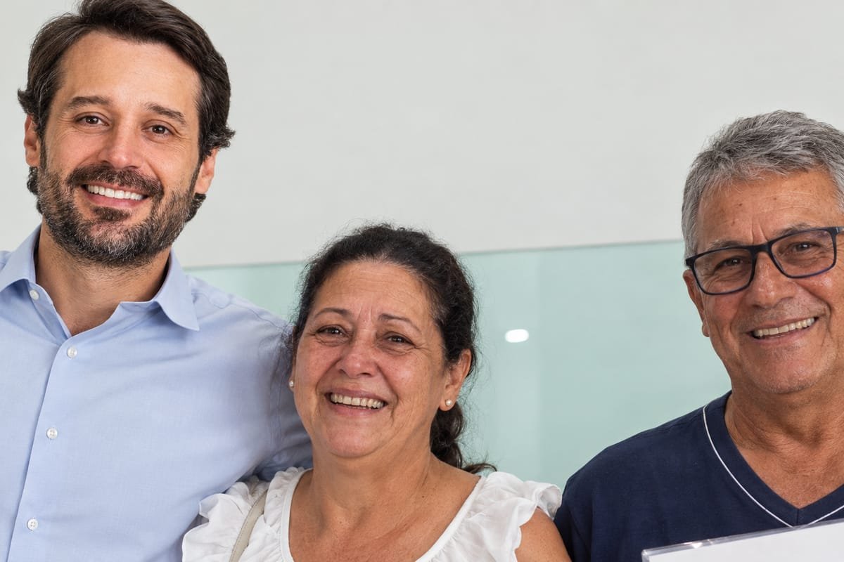Guilherme Piai, de camisa social azul clara, sorri abraçado a uma mulher de blusa branca e a um homem de óculos que segura um documento, em ambiente interno